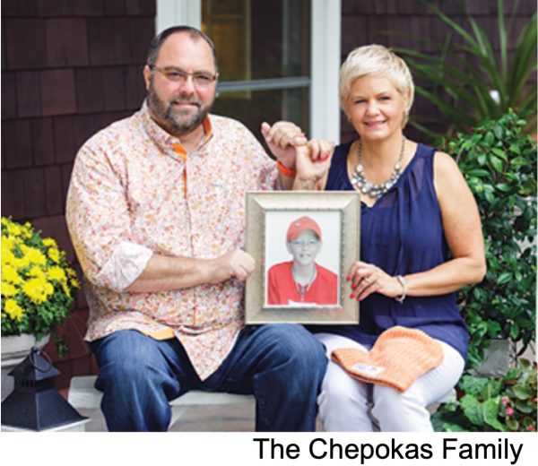 A man in a peach flowered shirt sits on the left of a woman with short white hair. They hold a picture of their son wearing a red shirt and baseball hat. Over the picture they hook pinkies.